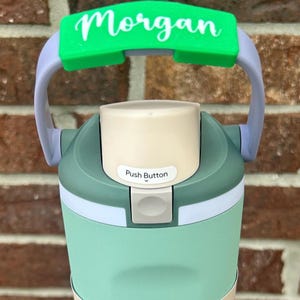 Op de afbeelding: Een turquoise en grijze waterfles met een groene handgreep met de naam "Morgan" in witte letters. De fles heeft een drukknop en een "FreeSip" tuit.