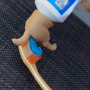 May include: A toothbrush with orange bristles and a blue toothpaste being dispensed from a tube. The toothpaste tube is white and blue. A novelty item shaped like a hand is dispensing the toothpaste.