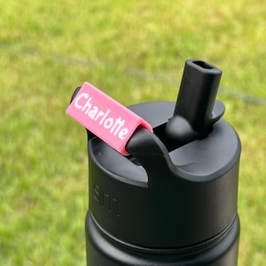May include: A black water bottle with a pink personalized name tag that says "Charlotte". The bottle has a black lid with a flip-top spout.