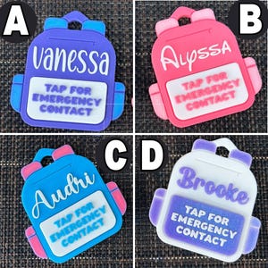 May include: Four colorful backpack-shaped tags. Each tag features a name and the text "TAP FOR EMERGENCY CONTACT." The tags are in purple, pink, blue, and white, with accents in contrasting colors. The names on the tags are Vanessa, Alyssa, Audri, and Brooke.