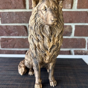 May include: A bronze-colored figurine of a collie dog in a seated position. The sculpture features detailed, textured fur patterns across the body, pointed ears, and a focused facial expression, finished with a metallic sheen and dark shading.