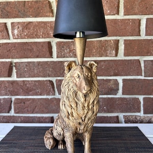 May include: A table lamp featuring a sculpted bronze-colored collie dog base with detailed fur texture. The lamp has a conical metal neck and a simple black tapered drum shade positioned directly above the dog's head, creating a functional light fixture.