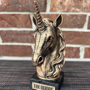 May include: A bronze-colored unicorn statue with a detailed horn, mane, and facial features. The statue is mounted on a black base with the name "THEODORE" engraved on a small plaque. The background features a brick wall.
