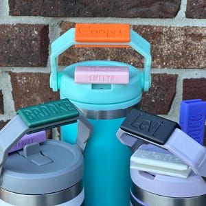 May include: Three stainless steel water bottles with personalized labels. The labels are in different colors and fonts. The bottles are in various shades of blue, purple, and gray.