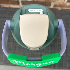Op de afbeelding: Een groene en beige tuitbeker met een groene handgreep en de naam "Morgan" in het wit. De beker heeft een wit deksel met de labels "Unlock" en "Lock". De handgreep is paars en groen.
