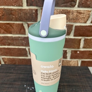 Op de afbeelding: Een mintgroene Owala waterfles met een lichtpaarse handgreep en een groene sluiting. De fles heeft een witte band en een beige deksel. De tekst op de fles luidt: "Hello friend. I'm leak proof. I'm cup holder friendly. And I have a built-in straw."