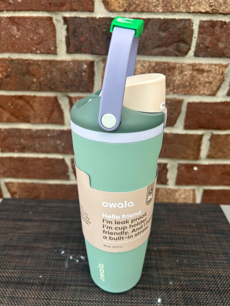 Op de afbeelding: Een mintgroene Owala waterfles met een lichtpaarse handgreep en een groene sluiting. De fles heeft een witte band en een beige deksel. De tekst op de fles luidt: "Hello friend. I'm leak proof. I'm cup holder friendly. And I have a built-in straw."