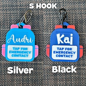 May include: Two blue backpack-shaped keychains with pink and red accents. One keychain says "Audri" and the other says "Kai". Both have the text "TAP FOR EMERGENCY CONTACT". One has a silver hook, the other a black hook.