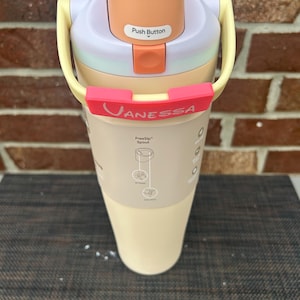 Op de afbeelding: Een veelkleurige waterfles met een drukknopdeksel. De fles heeft een lichtperzikkleurig deksel, een lichtpaarse bovenkant en een lichtgele handgreep. De naam "Vanessa" staat op een rode band rond de fles.