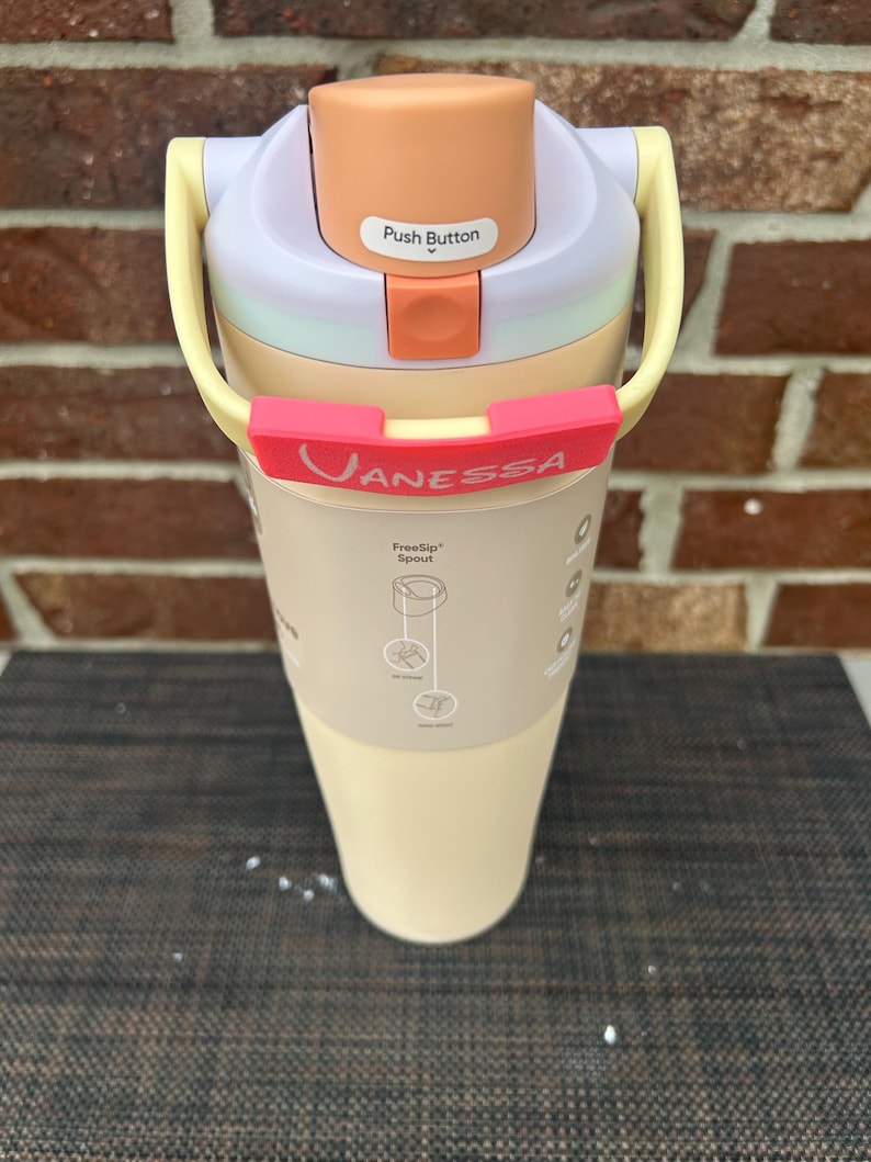 Op de afbeelding: Een veelkleurige waterfles met een drukknopdeksel. De fles heeft een lichtperzikkleurig deksel, een lichtpaarse bovenkant en een lichtgele handgreep. De naam "Vanessa" staat op een rode band rond de fles.