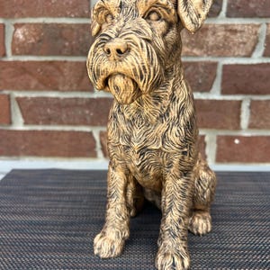May include: A detailed, seated dog statue with a textured, golden-brown finish. The sculpture features a long snout, bushy eyebrows, and a full beard. The dog's fur is intricately carved, adding depth and realism to the piece.