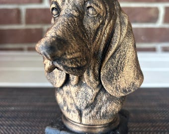 May include: A bronze-colored dog bust with a textured finish, mounted on a black rectangular base. The name "NIKKI" is displayed on a gold-colored plaque. The sculpture is set against a blurred brick wall background.