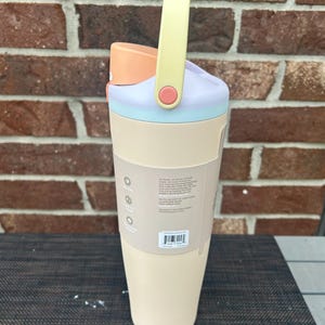 Op de afbeelding: Een pastelkleurige waterfles met een handvat. De fles heeft een lichtbeige basis, een lichtblauwe band, een lavendelkleurige bovenkant en een perzikkleurig deksel. Het handvat is geel met een roze accent. De fles heeft een label met tekst.