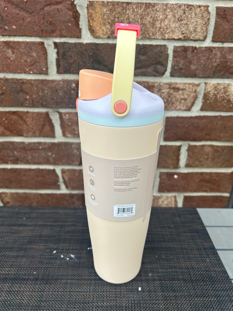 Op de afbeelding: Een pastelkleurige waterfles met een handvat. De fles heeft een lichtbeige basis, een lichtblauwe band, een lavendelkleurige bovenkant en een perzikkleurig deksel. Het handvat is geel met een roze accent. De fles heeft een label met tekst.