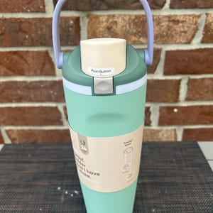 Op de afbeelding: Een mintgroene waterfles met een witte drukknopdeksel en een groene handgreep. De fles heeft een lichtbruine band met tekst en een diagram. De handgreep is groen en grijs. De fles staat op een donkere ondergrond.