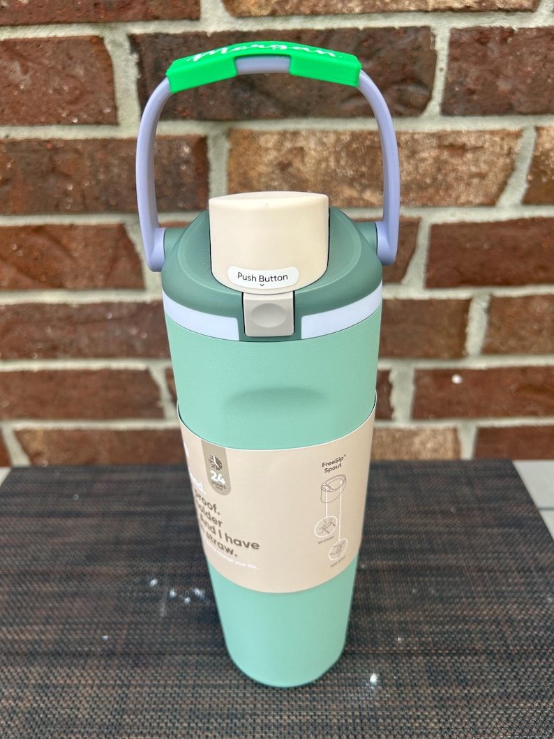Op de afbeelding: Een mintgroene waterfles met een witte drukknopdeksel en een groene handgreep. De fles heeft een lichtbruine band met tekst en een diagram. De handgreep is groen en grijs. De fles staat op een donkere ondergrond.