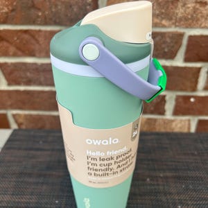 Op de afbeelding: Een mintgroene Owala waterfles met een lichtpaarse handgreep en een cr&egrave;mekleurig deksel. De fles heeft een ingebouwd rietje en de tekst "Hello friend. I'm leak proof. I'm cup holder friendly. And a built-in straw."