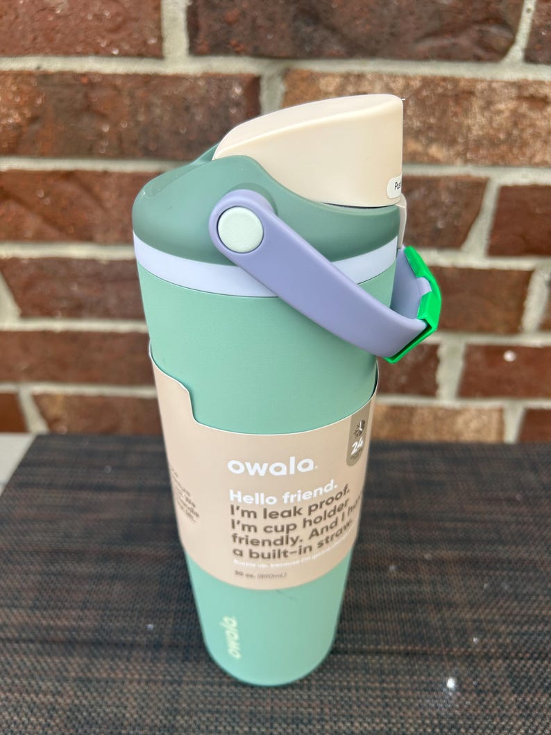 Op de afbeelding: Een mintgroene Owala waterfles met een lichtpaarse handgreep en een cr&egrave;mekleurig deksel. De fles heeft een ingebouwd rietje en de tekst "Hello friend. I'm leak proof. I'm cup holder friendly. And a built-in straw."