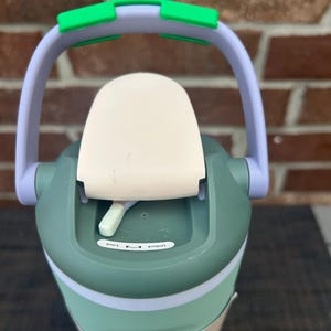 Op de afbeelding: Een groene en witte waterfles met een handvat en een klapdeksel. Het handvat is lichtpaars met groene accenten. De deksel heeft een witte bovenkant met een vergrendelingsmechanisme met de tekst "Lock" en "Unlock".