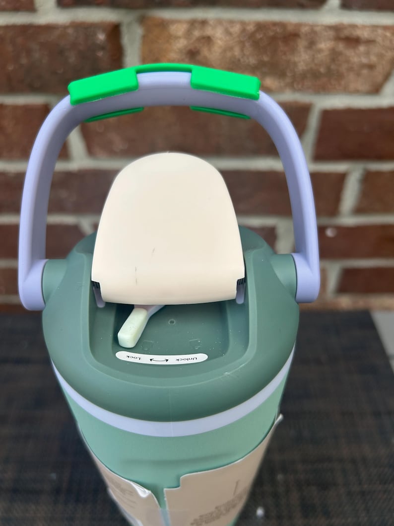 Op de afbeelding: Een groene en witte waterfles met een handvat en een klapdeksel. Het handvat is lichtpaars met groene accenten. De deksel heeft een witte bovenkant met een vergrendelingsmechanisme met de tekst "Lock" en "Unlock".