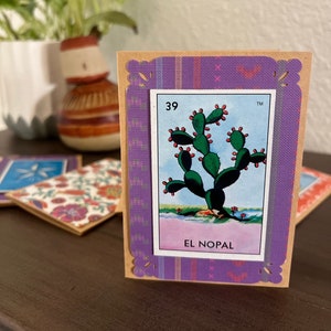 May include: A vintage-style card with a purple border and a brown background. The card features a colorful illustration of a prickly pear cactus with the text "39 EL NOPAL" printed on it.