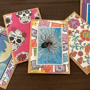 Könnte beinhalten: Ein Set aus sechs bunten Karten mit verschiedenen Designs. Die Karten zeigen Bilder von einem Schädel, einer Spinne, einem Mond, einem Stern und Blumenmustern. Die Karten sind mit bunten Papierschnipseln verziert und haben Nummern darauf. Die Karten sind aus Pappe und haben einen braunen Hintergrund.