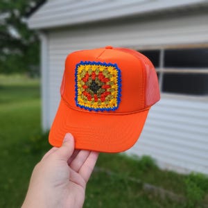 May include: A bright orange trucker hat featuring a square crochet patch on the front panel. The patch is composed of concentric layers in black, olive green, red, and yellow, finished with a vibrant blue scalloped crochet border edge.