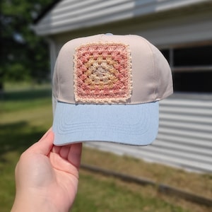 May include: A baseball cap featuring a light beige crown and a light blue denim brim. The front panel is adorned with a square, multi-colored crochet granny square patch in shades of pink, peach, and cream with a scalloped edge border.