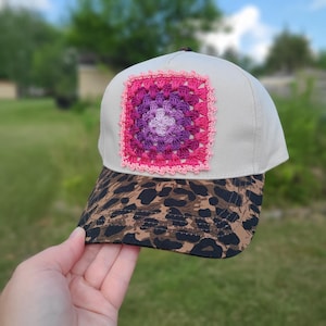 May include: A baseball cap with a cream-colored crown and a brown and black leopard print brim. The front of the crown features a square, multi-colored crochet patch in shades of light purple, dark purple, magenta, and a light pink scalloped border.