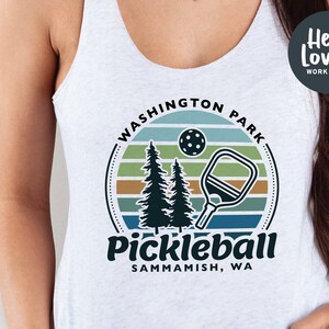 Może przedstawiać: Biała koszulka na ramiączkach z grafiką przedstawiającą zach&oacute;d słońca, dwa sosny, rakietę do pickleballa i tekst "Washington Park Pickleball, Sammamish, WA".
