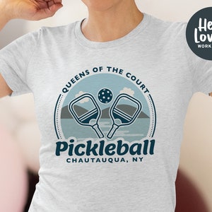 Może przedstawiać: Szara koszulka z niebiesko-białym nadrukiem graficznym z napisem "Queens of the Court Pickleball Chautauqua, NY". Grafika przedstawia dwie rakiety do pickleballa i piłkę.