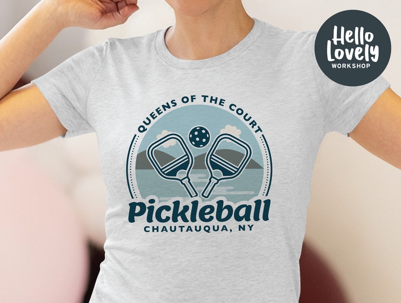 Może przedstawiać: Szara koszulka z niebiesko-białym nadrukiem graficznym z napisem "Queens of the Court Pickleball Chautauqua, NY". Grafika przedstawia dwie rakiety do pickleballa i piłkę.