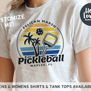 Może przedstawiać: Biały T-shirt z grafiką przedstawiającą rakietę do pickleballa i tekstem "Pelican Marsh Pickleball Naples, FL". Tekst "Customize Me!" znajduje się w lewym g&oacute;rnym rogu, a "Hello Lovely Workshop" w prawym g&oacute;rnym rogu.