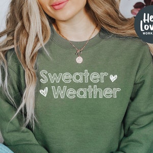 May include: An olive green sweatshirt with the words "Sweater Weather" in white, handwritten-style lettering. Two small white hearts are on either side of the word "Weather". The sweatshirt has a crew neck and long sleeves. The image also includes the text "Hello Lovely Workshop".