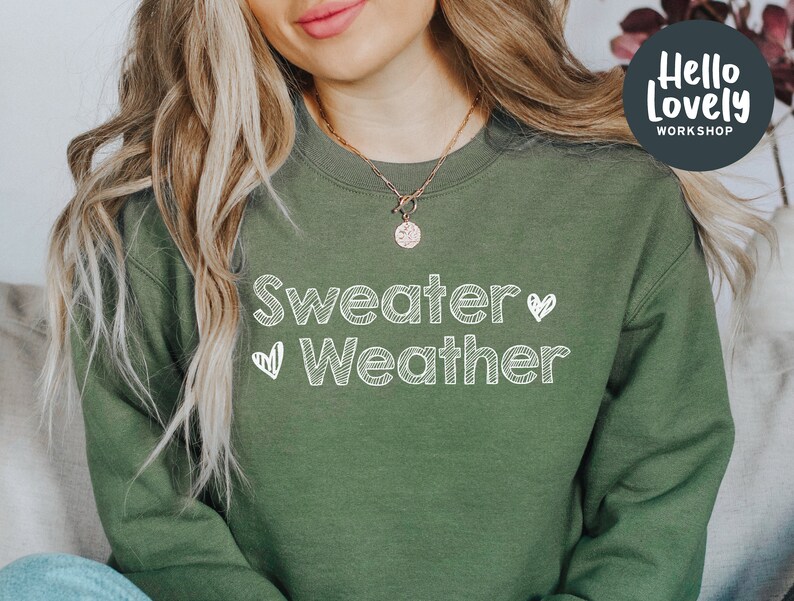 May include: An olive green sweatshirt with the words "Sweater Weather" in white, handwritten-style lettering. Two small white hearts are on either side of the word "Weather". The sweatshirt has a crew neck and long sleeves. The image also includes the text "Hello Lovely Workshop".