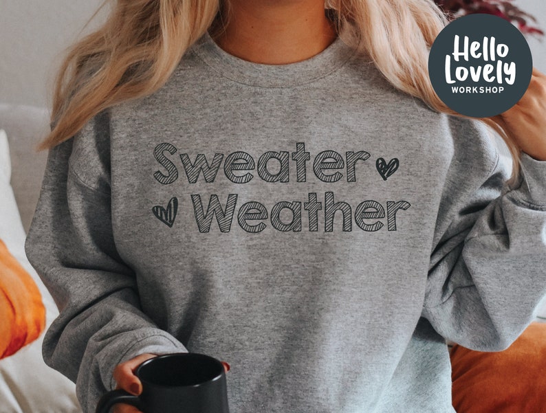 May include: A heather gray sweatshirt with the words "Sweater Weather" in black, outlined text, and two small heart graphics. The sweatshirt is part of the Hello Lovely Workshop collection.