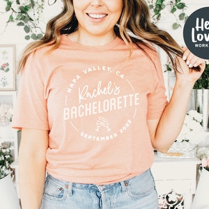 Puede incluir: Una camiseta de color melocot&oacute;n con un gr&aacute;fico circular blanco que dice "Napa Valley, CA Rachel's Bachelorette Septiembre 2022" con un gr&aacute;fico de dos copas de champ&aacute;n.