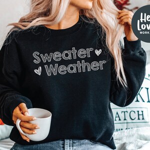 May include: Black sweatshirt with the text "Sweater Weather" in white script, with two small heart graphics. The sweatshirt is a crew neck style. A white mug is held in the person's hand. The Hello Lovely Workshop logo is in the upper right corner.