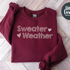 May include: A maroon sweatshirt with the words "Sweater Weather" in white, handwritten-style font. A small heart is next to the word "Weather". The sweatshirt has a crew neck and long sleeves. The brand name "Hello Lovely Workshop" is in a circle in the upper right corner.