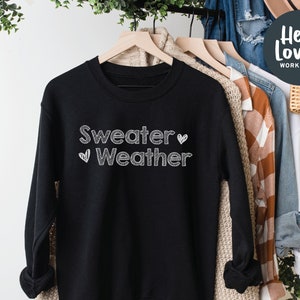 May include: Black sweatshirt with the text "Sweater Weather" in white, handwritten-style lettering. A white heart graphic is next to the word "Weather". The sweatshirt is hanging on a wooden hanger with other clothing items in the background.