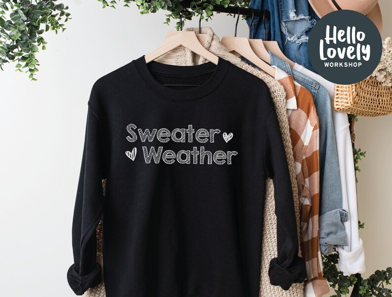 May include: Black sweatshirt with the text "Sweater Weather" in white, handwritten-style lettering. A white heart graphic is next to the word "Weather". The sweatshirt is hanging on a wooden hanger with other clothing items in the background.