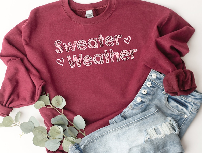 May include: A maroon sweatshirt with the words "Sweater Weather" in white script, accented with two small hearts. The sweatshirt is paired with denim shorts. A sprig of greenery is placed near the clothing.