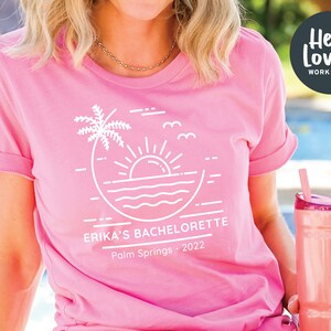 Peut inclure: T-shirt rose avec un graphique blanc d'un palmier, du soleil et des vagues. Le texte "Erika's Bachelorette Palm Springs - 2022" est imprim&eacute; sur le t-shirt.