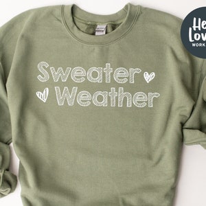 May include: An olive green sweatshirt with the words "Sweater Weather" in white, handwritten-style font, with a heart symbol. The sweatshirt has a crew neck and long sleeves. A circular logo with the text "Hello Lovely Workshop" is in the upper right corner.