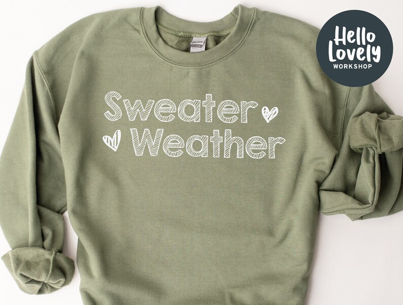 May include: An olive green sweatshirt with the words "Sweater Weather" in white, handwritten-style font, with a heart symbol. The sweatshirt has a crew neck and long sleeves. A circular logo with the text "Hello Lovely Workshop" is in the upper right corner.