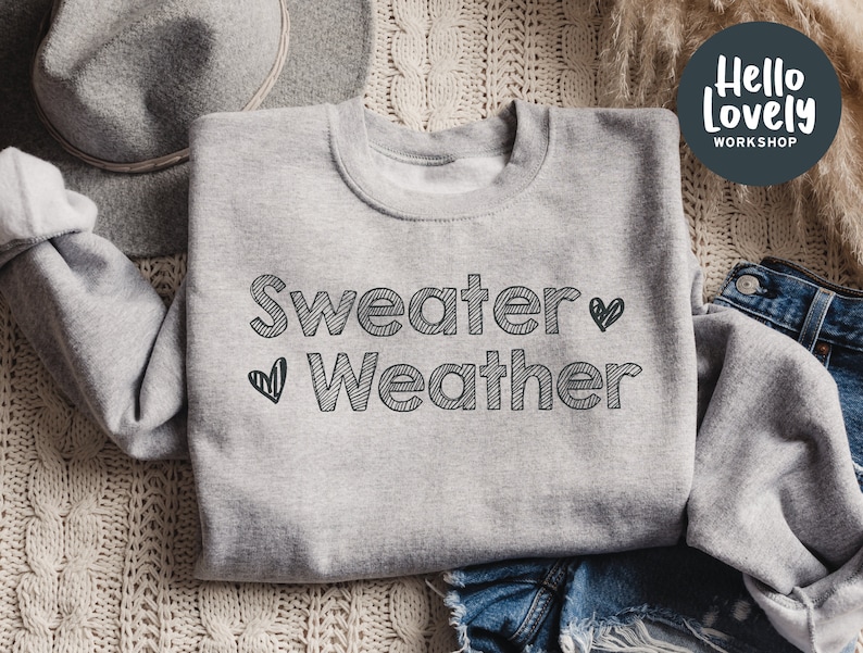 May include: A light gray sweatshirt with the words "Sweater Weather" in black, outlined text and two black heart graphics. The sweatshirt is folded on a cream-colored knitted surface, with a gray hat and denim shorts visible.