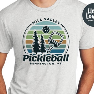 Może przedstawiać: Szary T-shirt z retro grafiką przedstawiającą zach&oacute;d słońca, dwa sosny, rakietę do pickleballa i piłkę do pickleballa. Na koszulce nadrukowany jest tekst "Mill Valley Pickleball Bennington, VT".