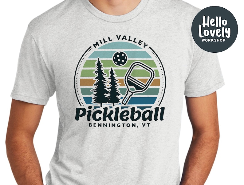 Może przedstawiać: Szary T-shirt z retro grafiką przedstawiającą zach&oacute;d słońca, dwa sosny, rakietę do pickleballa i piłkę do pickleballa. Na koszulce nadrukowany jest tekst "Mill Valley Pickleball Bennington, VT".