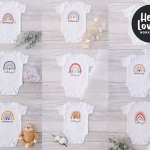 Può includere: Nove body bianchi per neonati con un arcobaleno e il testo "birthday girl" stampato sul davanti. I body sono disposti in tre file di tre. I body hanno diversi disegni e colori di arcobaleno.