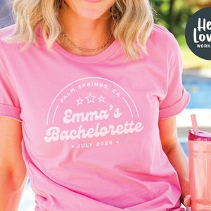 Puede incluir: Camiseta rosa con un gr&aacute;fico blanco de estilo retro que dice "Palm Springs, CA" con tres estrellas y "Emma's Bachelorette" con la fecha "Julio 2022".
